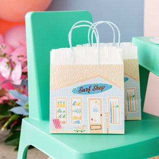 Two Surf's Up Party Bags by Daydream Society are set on a green chair, with flowers blooming in the background.