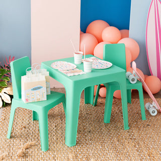 A small turquoise table with two matching chairs is arranged for a festive farewell, showcasing the Surf's Up Party Bag from Daydream Society filled with party favors, alongside drinks and plates. In the background, a pink skateboard adds to the ambiance, perfectly capturing the essence of beachside surf culture.