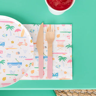 A wooden knife and fork are placed on a colorful paper napkin featuring tropical designs, next to the Surf's Up Small Plates from Daydream Society and a cup of red liquid on the green table. This arrangement is part of the vibrant Surf's Up Collection, perfectly capturing the essence of beachside surf culture for your dining experience.