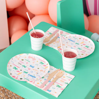 Two place settings on a green table feature patterned plates from Daydream Society's Surf's Up Small Plates collection, accompanied by wooden cutlery and striped cups filled with a red drink. Pink balloons in the background add a splash of fun reminiscent of beachside surf culture.