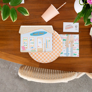 Top view of a table adorned with decorative paper items, including Daydream Society's Surf's Up Surf Shop Large Plates that echo beachside surf culture, alongside patterned paper, near a wicker chair and potted plant.