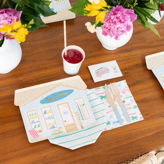 The table setting encapsulates beachside surf culture with Daydream Society's Surf's Up Surf Shop Large Plates, a wooden fork and knife, a cup with a straw, vibrant flowers in vases, and a small card featuring "Surf's Up" along with an illustration of a van.