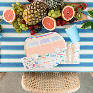 The Surf's Up Collection stands out with a striped tablecloth featuring Daydream Society's Surf's Up Surf Van Large Plates, a vibrant fruit arrangement with pineapple, grapes, and grapefruit, and a blue cup. The partially visible wicker chair adds a touch of beachside surf culture.