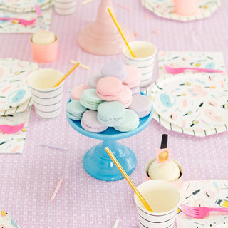A pastel-colored dessert table set for a sweet slumber party with a stand of multicolored macarons and coordinating paper cups with straws, all on a playful Daydream Society tablecloth containing Sweet Dreams Large Napkins.