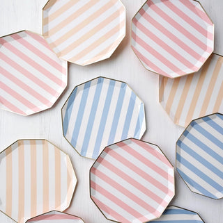 An assortment of octagonal Sweet Peach Signature Cabana Stripe paper plates with pastel pink and blue diagonal stripes arranged in a scattered pattern on a light wooden surface, suggesting a festive or celebratory occasion.