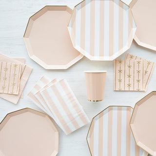 A collection of Sweet Peach Signature Cabana Stripe Plates by Bonjour Fête arranged neatly on a light wooden surface, featuring octagonal plates, cups, and napkins with a gold motif.