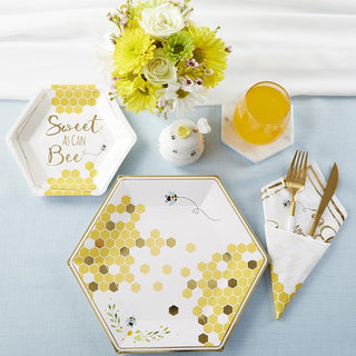 Hexagonal plates and napkins with a **Sweet as Can Bee Cocktail Plate** design by **Kate Aspen**, a floral bouquet, a honey pot, a glass of orange juice, and gold-colored cutlery on a light blue tablecloth create the perfect setting for your baby shower.