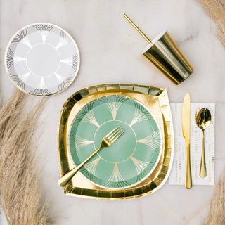 Create an elegant table setting with Jollity & Co's Gatz Collection gold-rimmed dinner plates, paired with a fork resting on a sage green plate. Incorporate gold utensils placed on a guest napkin and finish the look with a gold cup set on a pristine white surface, complemented by decorative pampas grass.