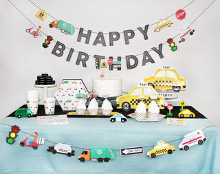 A transportation-themed birthday party setup featuring Merrilulu's Transportation Garland with 14 vibrant pennants, including a "Happy Birthday" banner, alongside paper cups and plates adorned with vehicles like taxis, police cars, and ambulances on a decorated table.