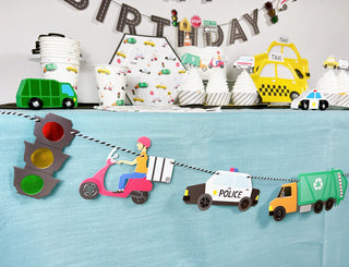 The birthday party table was adorned with Merrilulu Transportation-themed items, including a vibrant Transportation Garland featuring a scooter, police car, and recycling truck. Various related paper cups and plates completed the festive look for an unforgettable vehicle-inspired celebration.