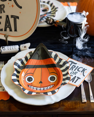 The Halloween-themed table setting includes a pumpkin-faced plate with a witch hat design, "Trick or Treat" cocktail napkins by My Mind’s Eye, a spooky black goblet emitting smoke, and festive décor perfect for any Halloween party.