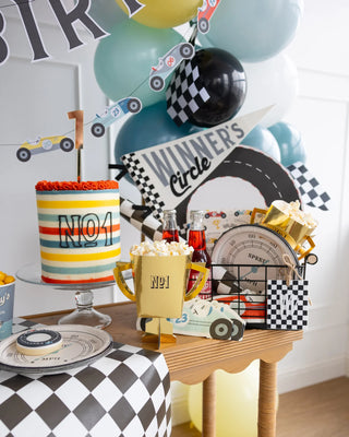 Birthday party setup featuring a race car theme with a striped cake, gold My Mind’s Eye Trophy Treat Cups filled with popcorn, and black-and-white checkered decorations, including balloons and race track props.