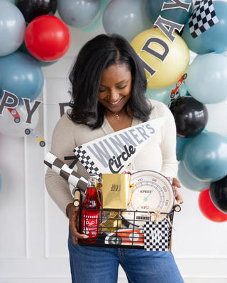 A woman holds a gift basket themed with a racing motif, featuring a "Winner's Circle" sign, My Mind’s Eye Trophy Treat Cups, a red soda bottle, snacks, and other small items. A "Happy Birthday" balloon garland hangs in the background, perfectly capturing the excitement of a car-themed birthday party.