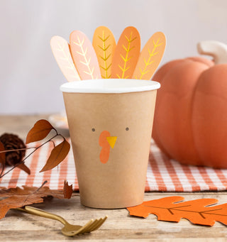 A Turkey Paper Cup by My Mind’s Eye, designed to resemble a turkey with feather cutouts at the top, sits on a wooden table adorned with autumn leaves, a pumpkin, and a fork nearby—perfect for delighting Thanksgiving guests during your holiday gathering.