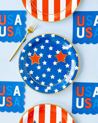 A patriotic table setting featuring star-spangled blue plates, coordinating red and white striped plates, a golden fork, "USA" banners, and My Mind's Eye USA Scallop Paper Cocktail Napkins, all arranged on a