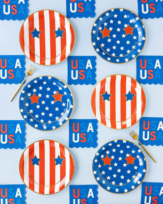 A festive table setting featuring patriotic My Mind’s Eye Star & Stripe Smiley Paper Plate Set with red and white stripes, blue borders, and white stars on a tablecloth adorned with "USA" motifs, ready for a fun celebration.
