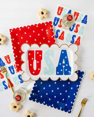 A patriotic table setting with "USA" themed decorations in red, white, and blue, featuring star-shaped cookies, My Mind’s Eye USA Scallop Paper Plates, and napkins arranged for a festive celebration.