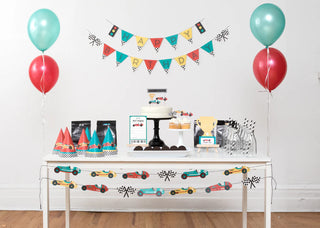 Table set up for a children's birthday party with a racing theme, featuring balloons, a cake, party hats, snacks, and a "Happy Birthday" banner along with Merrilulu's Vintage Race Car Party Garland.