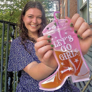 A smiling person holds an NPW Western Boot Shaped Drink Pouch with the text "LET'S GO GIRLS" in white letters—ideal for any cowgirl bachelorette party.