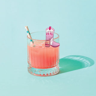 A glass with a pink drink, ice, a blue and white straw, and a pink and white cowboy boot garnish on a light blue background featuring playful NPW Western Drink Markers-6 Pack—a perfect novelty gift.