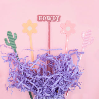 Five decorative NPW Western Drink Stirrers, featuring two cacti, two flowers, and one spelling out "Howdy," are displayed in a jar filled with purple shredded paper against a pink background.