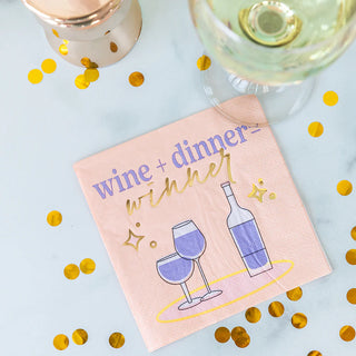 A playful Jollity & Co Witty Cocktail Napkin with the equation "wine + dinner = winner" rests on a surface scattered with gold confetti, next to a glass of white wine with a blurred candleholder in the background.