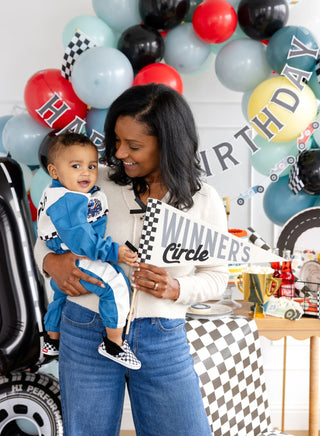 A smiling woman holds a young child dressed in a racing outfit at a racing-themed birthday party. They are surrounded by balloons, banners, and My Mind’s Eye Winners Circle Felt Pennant that declare the little one the birthday champ.