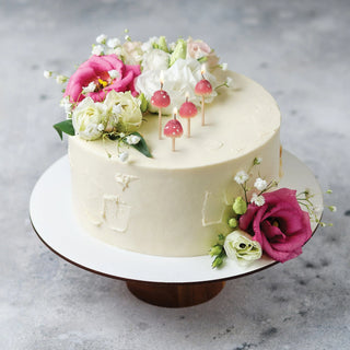 A white frosted cake on a stand, adorned with pink and white flowers and topped with three charming Mushroom Birthday Candles by Fred and Friends, adding an enchanting touch.