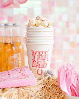 A stack of festive My Mind's Eye Yeehaw Paper Party Cups with "yeehaw" text, filled with popcorn, beside pink utensils and bottled drinks on a straw surface.