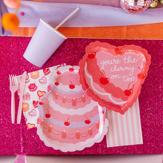 Party table setting featuring Jollity & Co's "You're the Cherry On Top" die-cut dessert plates, a heart-shaped card with the message "you're the cherry on top," a striped napkin, a white cup, plastic cutlery, and a glittery pink tablecloth highlighted with gold foil.