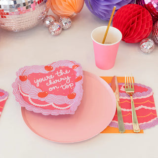 A table setting featuring the Jollity & Co "You're the Cherry On Top Dessert Plates," accompanied by a pink heart-shaped gift box, pink plate, and gold foil cutlery, exudes elegance. The ensemble includes a die-cut plate, a pink cup, a colorful napkin, and is surrounded by decorative orbs, creating a charming vintage Victorian ambiance.