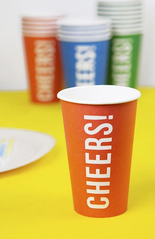 A close-up of an orange recyclable paper cup with the words "Cheers Red Paper Cups" from Talking Tables printed vertically. Blurred in the background are additional eco-friendly tableware cups in various colors.