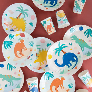 An assortment of colorful party plates and My Little Day Dino Jurassic Cups with playful prehistoric decorations scattered on a burgundy background, perfect for a child's birthday celebration.