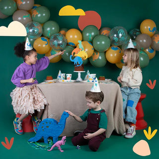 Two girls and a boy celebrate at a dinosaur-themed birthday party, with a balloon backdrop, festive hats, table settings, and toy dinosaurs. The table is adorned with My Little Day Dino Jurassic Cups among the pre.