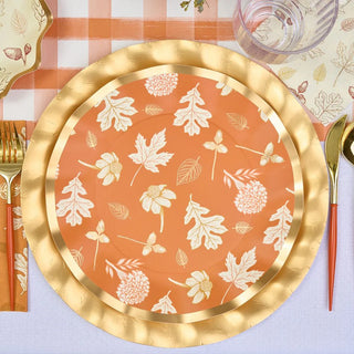 A round Sophistiplate Harvest Garden Dinner Plate, featuring gold foil edges and autumn-themed floral patterns on an orange backdrop, is placed on a checkered tablecloth with gold cutlery to the left and a glass to the top right—perfect for holiday gatherings.