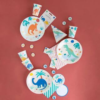 A colorful assortment of My Little Day's Dinosaur Jurassic Plates and cups with cheerful dinosaur illustrations, scattered playfully on a pink background, suggesting a prehistoric theme for a festive celebration.