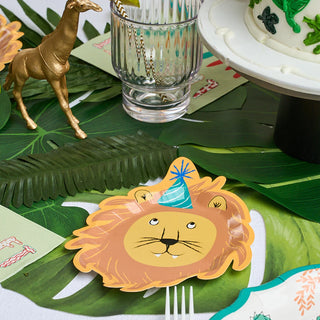 A holiday gathering table setting featuring the adorable Party Animal Lion Small Plate from Sophistiplate, showcasing a lion in a blue party hat, accompanied by a gold giraffe figurine, a glass with straw, and part of a cake adorned with green leaves.