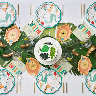 A table setting with jungle-themed decorations, including animal-shaped plates, leaf-shaped placemats, and various jungle motifs. Utensils, glasses, and holiday napkins featuring the "Party Animal Napkin" design by Sophistiplate are neatly arranged among the festive party tableware.