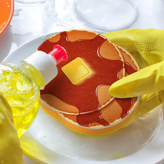 A person wearing yellow gloves washes a nostalgic diner plate using The Breakfast Scrub from Fred and Friends, a sponge designed to look like pancakes with syrup and a pat of butter, while applying dish soap.