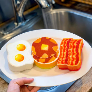 A hand holds a nostalgic diner plate featuring three kitchen sponges from Fred and Friends' The Breakfast Scrub set, designed to look like fried eggs, a pancake with syrup and butter, and bacon strips. These high-quality sponges are showcased against the backdrop of a kitchen sink with running water.