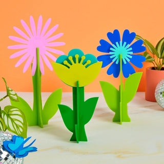 Three vibrant Kailo Chic die-cut acrylic flowers in pink, green, and blue stand cheerfully against an orange backdrop, evoking a playful and creative springtime garden vibe.