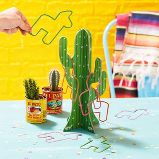 A hand placing a yellow airplane-shaped puzzle piece on a Ginger Ray Hoopla Cactus Party Game stand, with small cacti and colorful confetti on a table.