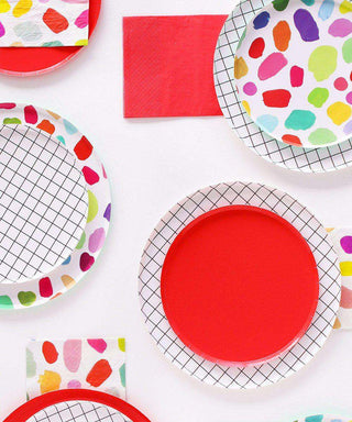 A vibrant arrangement of Oh Happy Day Kindah Plates - 7 inch with colorful patterns alongside red napkins on a white surface, suggesting a cheerful party or celebration setting.
