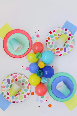 A festive overhead shot of colorful party supplies, including Oh Happy Day Kindah Plates - 7 inch, vibrant balloons, rolls of tape, and confetti on a white background, arranged in a playful and inviting composition.