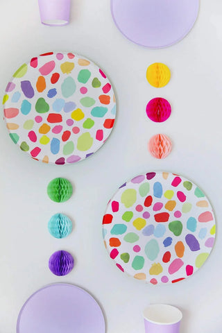A colorful party table setting with two Oh Happy Day Kindah Plates - 7 inch, coordinating paper cups, and a decorative arrangement of solid and honeycomb tissue paper balls on a white background.