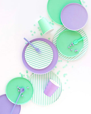 An assortment of mint green and purple party tableware, including Oh Happy Day Mint Stripe Plates - 7 inch, cups, and polka-dotted napkins, playfully arranged with scattered multicolored confetti.