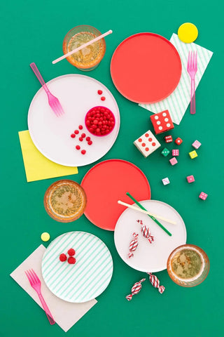 A vibrant tabletop arrangement featuring a colorful assortment of Oh Happy Day Mint Stripe Plates - 7 inch, utensils, and small bowls of food, accompanied by dice and candies, all set against a striking green background, evoking a playful vibe.