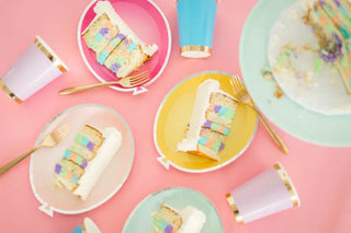 Pastel-colored unicorn themed party with slices of layered cake on paper plates, complemented by gold forks and Posh Lilac You Lots Cups with gold foiled rims, arranged on a pink tablecloth from the Jollity & Co Posh Collection.