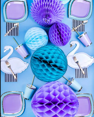 A vibrant party table setting featuring a cohesive blue and purple color palette, with elegant swan plates, assorted paper honeycomb decorations, striped napkins, Posh Lilac You Lots Cups with gold foiled rims from Jollity & Co.