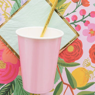 A Posh PinkAholic cup with a wooden stirrer against a vibrant floral background, flanked by green and gold geometric napkins.